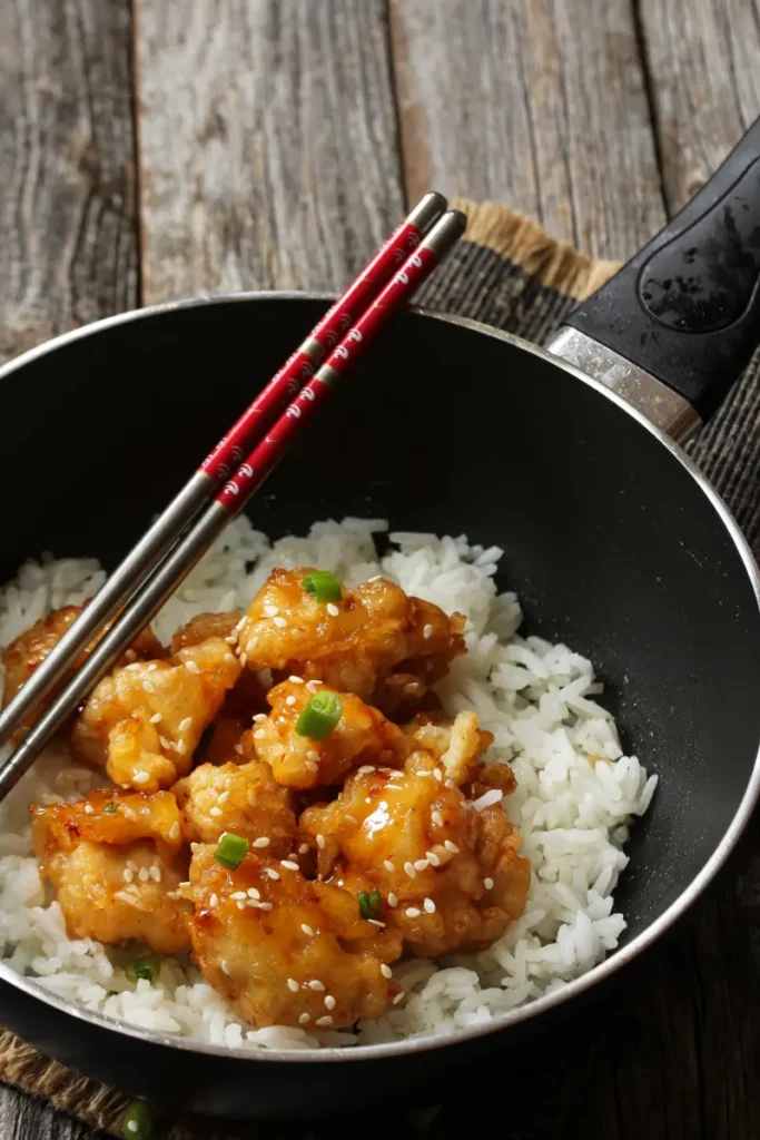 A bowl of crispy Honey Sesame Chicken on white rice, sprinkled with sesame seeds and green onions, with a pair of red chopsticks resting on top, set on a rustic wooden table.