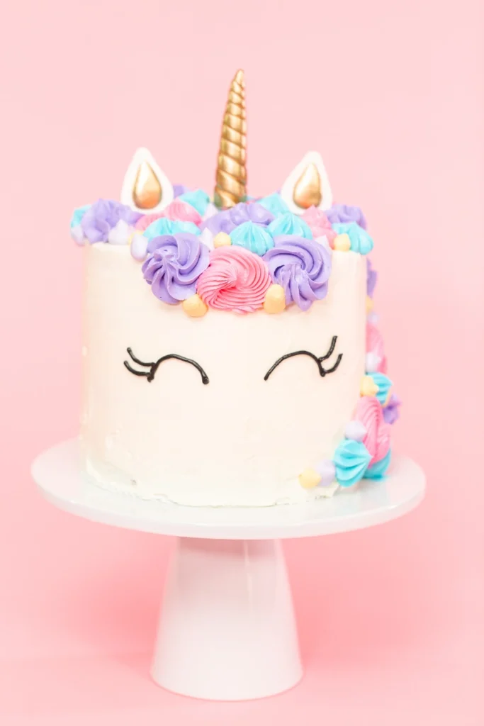 A unicorn-themed cake with a gold horn and white ears, topped with swirls of purple, pink, and blue icing, on a white cake stand against a pastel pink background.