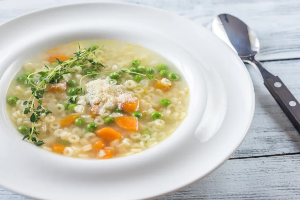 Bowl of pastina soup with carrots, peas, and a sprinkle of grated cheese, garnished with thyme, served with a spoon on the side.