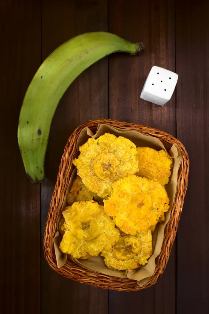 A wicker basket containing patacones beside a whole green plantain and a white six-sided dice, all on a dark wooden surface.