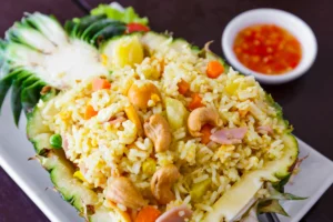 A plate of pineapple fried rice served in a hollowed-out pineapple half, garnished with green leaves, accompanied by a small bowl of red sauce on the side.