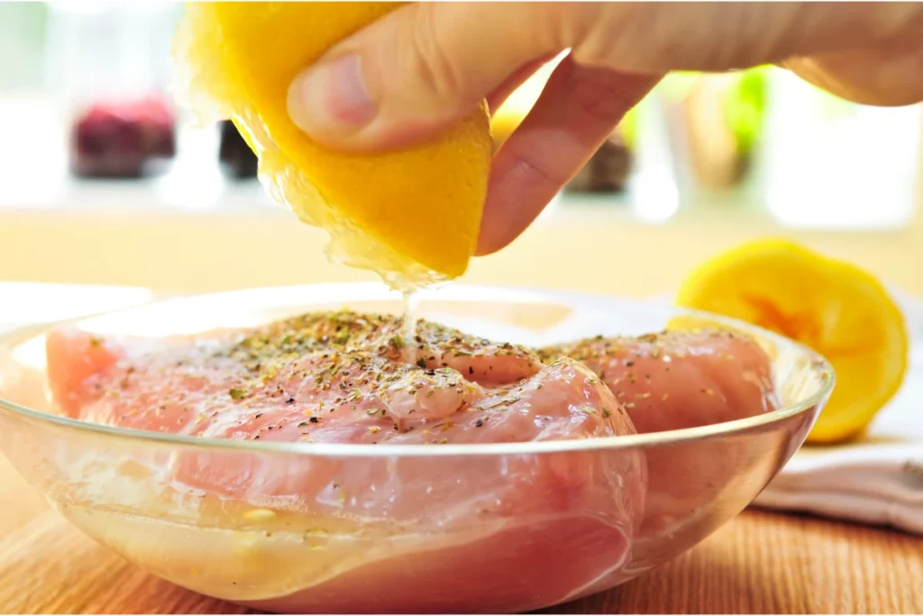 Hand squeezing lemon juice over seasoned raw chicken breasts in a glass bowl, preparing for a Greek marinade.