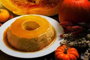 Pumpkin flan with a glossy caramel sauce on a white plate, surrounded by autumn decorations including a cut pumpkin, small gourds, and dried flowers.