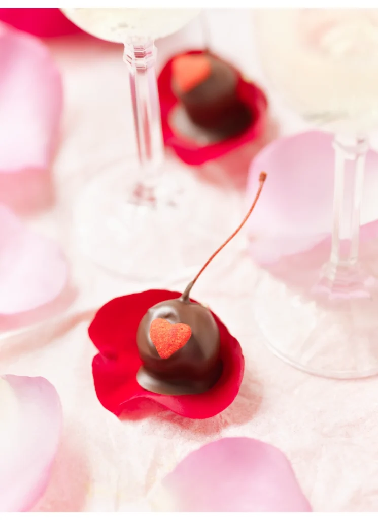 A chocolate-covered cherry adorned with a glittery heart, resting on a red rose petal, with soft focus on champagne flutes and rose petals in the background.