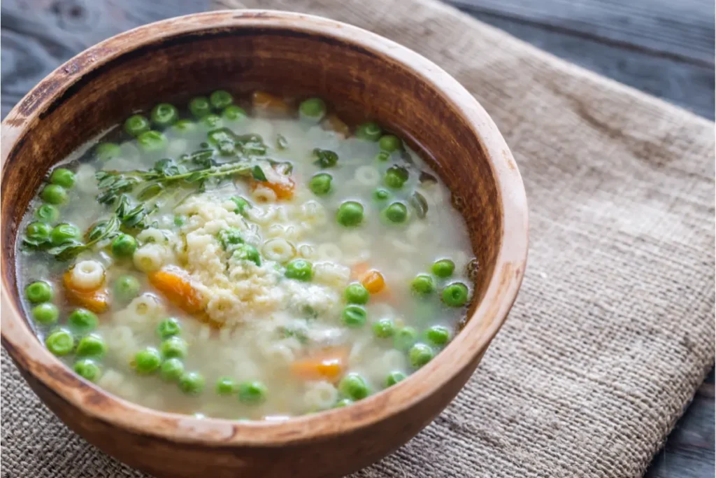 Rustic wooden bowl filled with pastina soup, featuring green peas, carrot slices, and garnished with herbs, on a burlap-covered table.