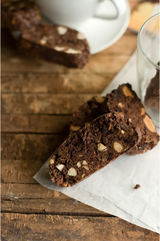 Chocolate biscotti with nuts on parchment, with blurred white mug and glass in background, atop a wooden table.