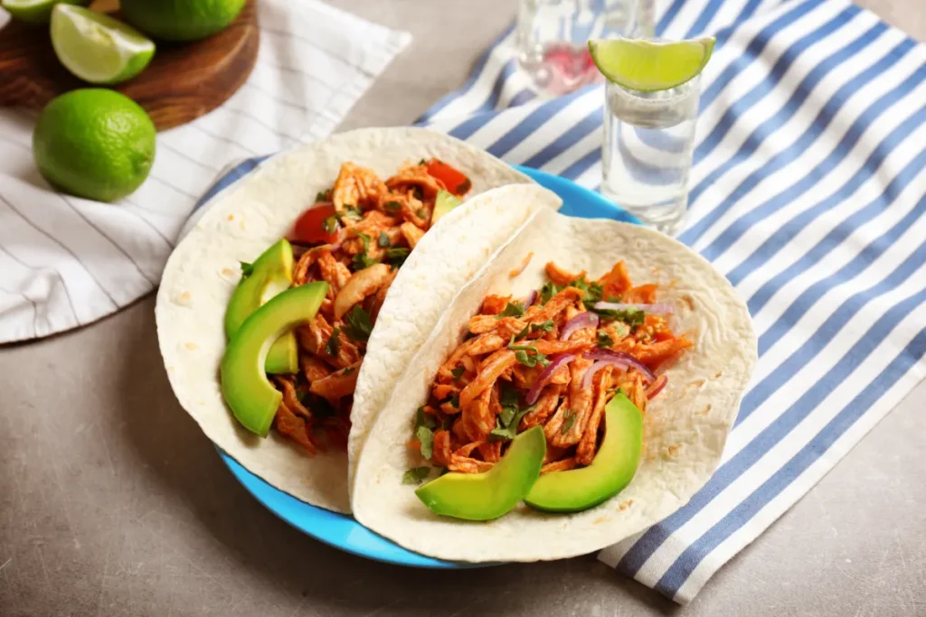 Two chicken tacos on a blue-rimmed plate with slices of avocado, topped with onions and cilantro. A shot glass of tequila and lime wedges are visible in the background, all set on a striped blue and white tablecloth.