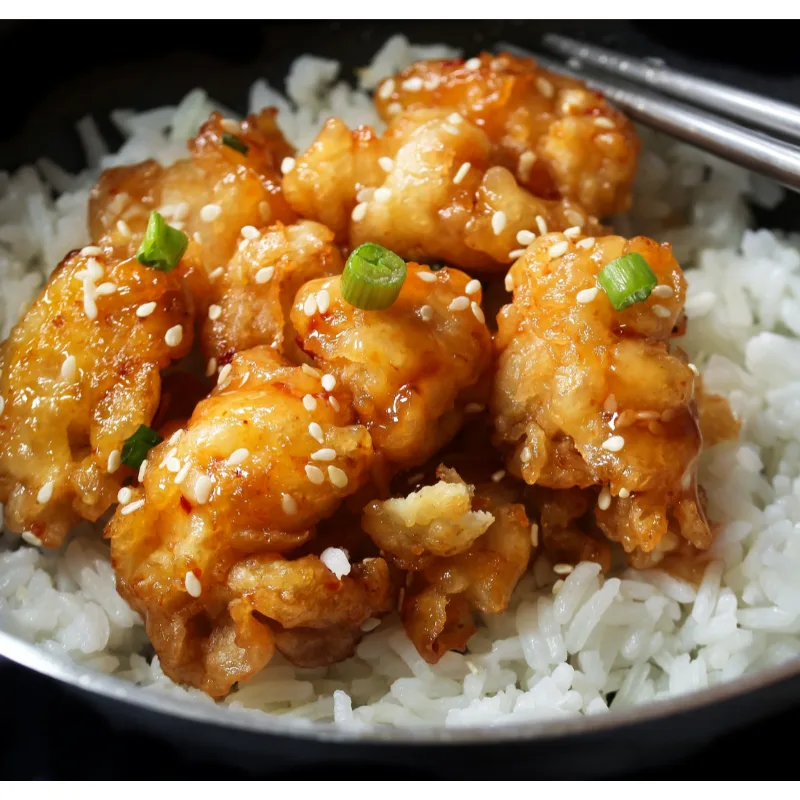 Close-up of Panda Express Honey Sesame Chicken, with crispy chunks glazed in honey sauce and sprinkled with sesame seeds, served over a bed of white rice, topped with green onions, in a black bowl with chopsticks.