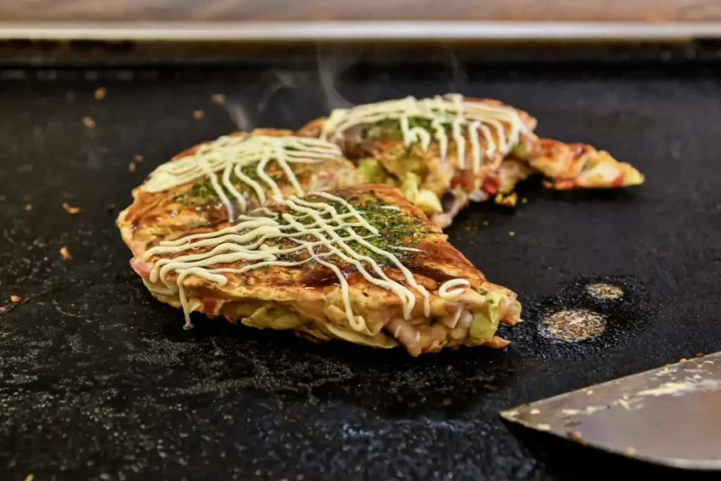 Two pieces of okonomiyaki with mayonnaise and green seasoning on a griddle, steam rising, next to a spatula.