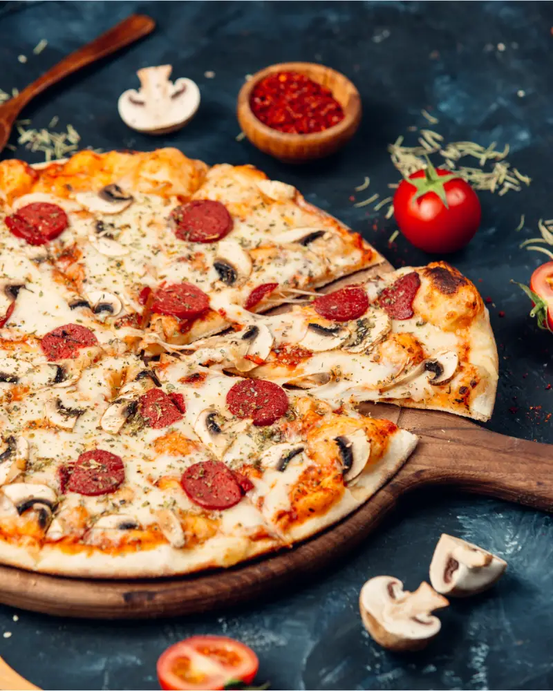 A thin crust pizza with pepperoni, mushrooms, and melted cheese on a wooden pizza paddle, with a tomato, a bowl of chili flakes, and fresh herbs in the background on a dark textured surface.