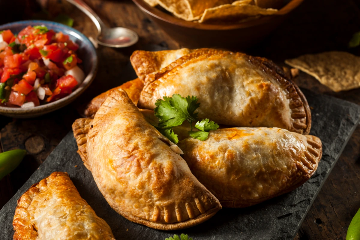 Three golden-brown pumpkin empanadas on a slate surface, accompanied by a bowl of tomato salsa and tortilla chips in the background.