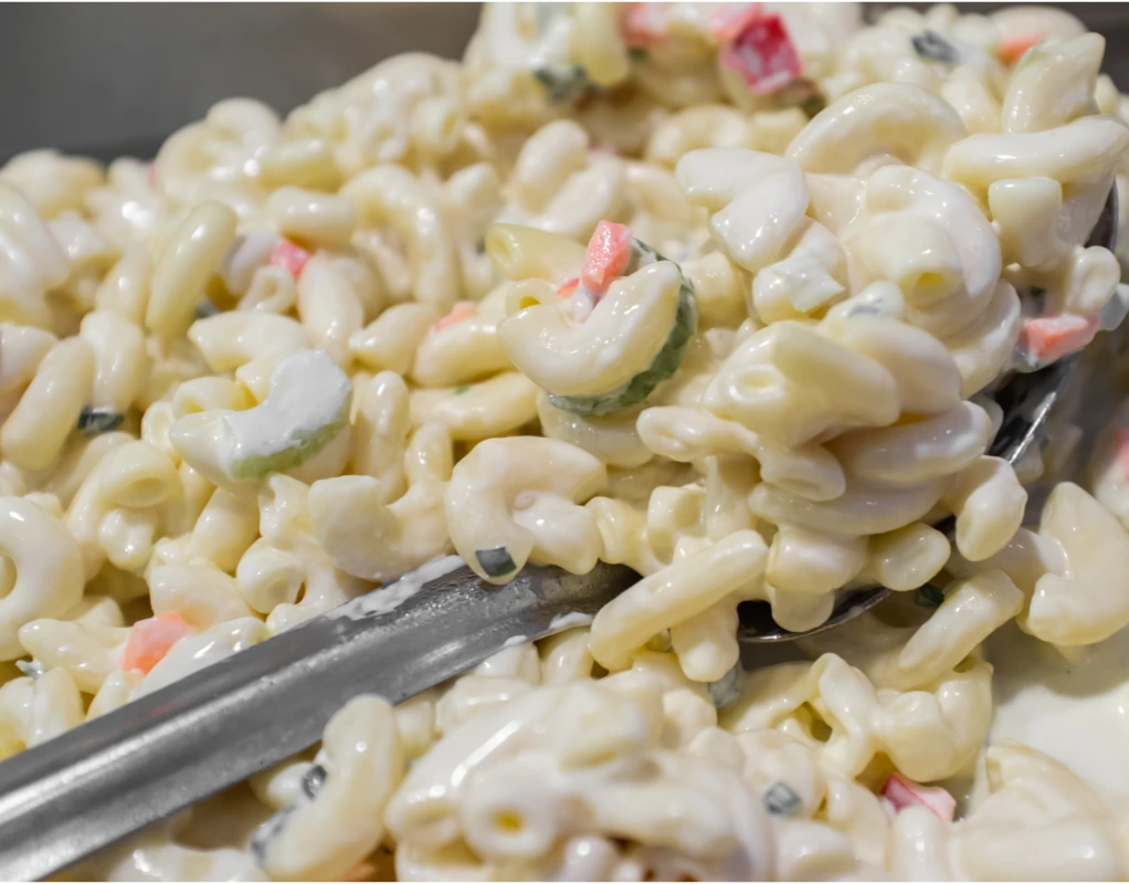 Scooping Hawaiian Macaroni Salad with a fork, showing the glossy, creamy dressing coating the pasta, mixed with green onions and bits of red bell pepper.