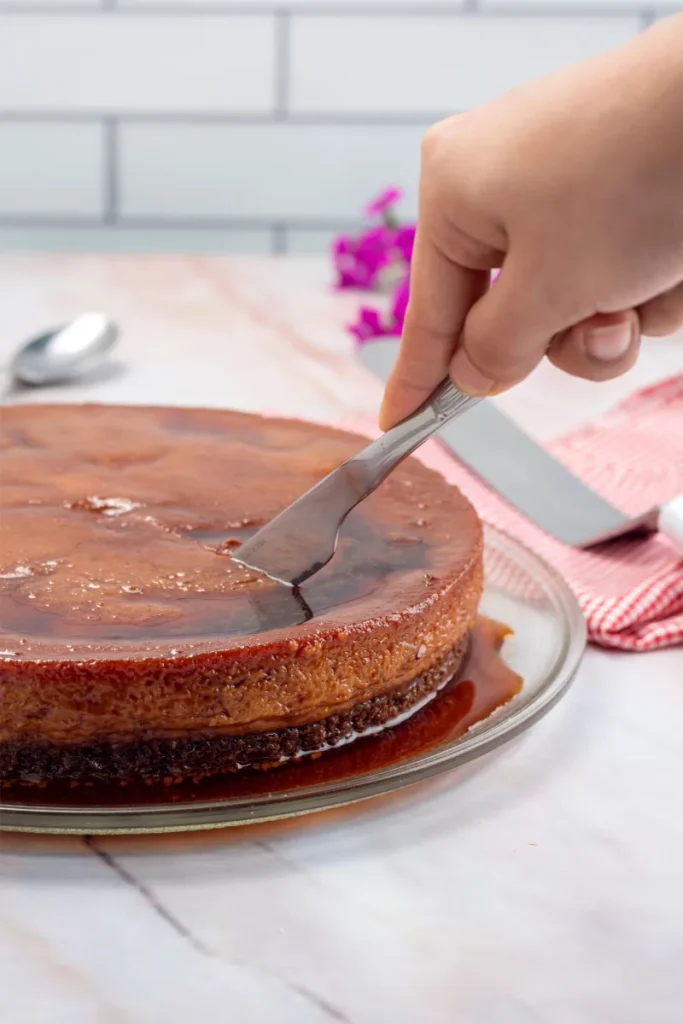 A hand is cutting into a chocolate flan on a glass plate with a metal knife, indicating the beginning of serving. The dessert is set on a marble countertop with a pink striped napkin and purple flowers in the soft-focused background.