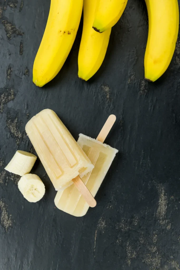 Two banana popsicles on a dark slate surface with whole bananas in the background and a banana slice in the foreground.