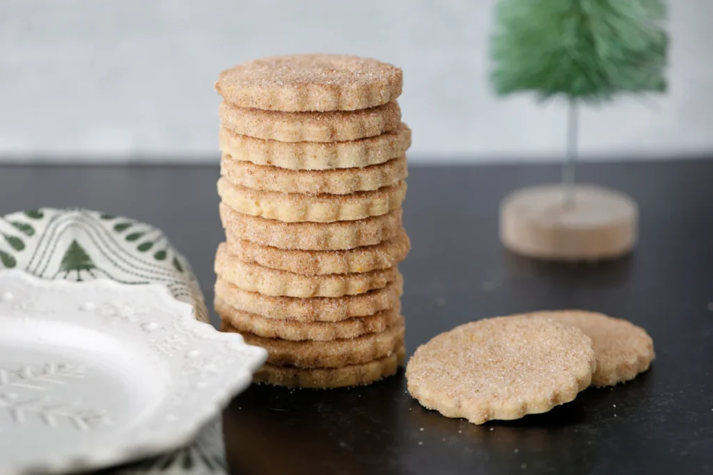 Alt Text: "A neat stack of round biscochitos cookies, dusted with cinnamon sugar, next to a decorative plate and a small green tree ornament on a dark countertop.