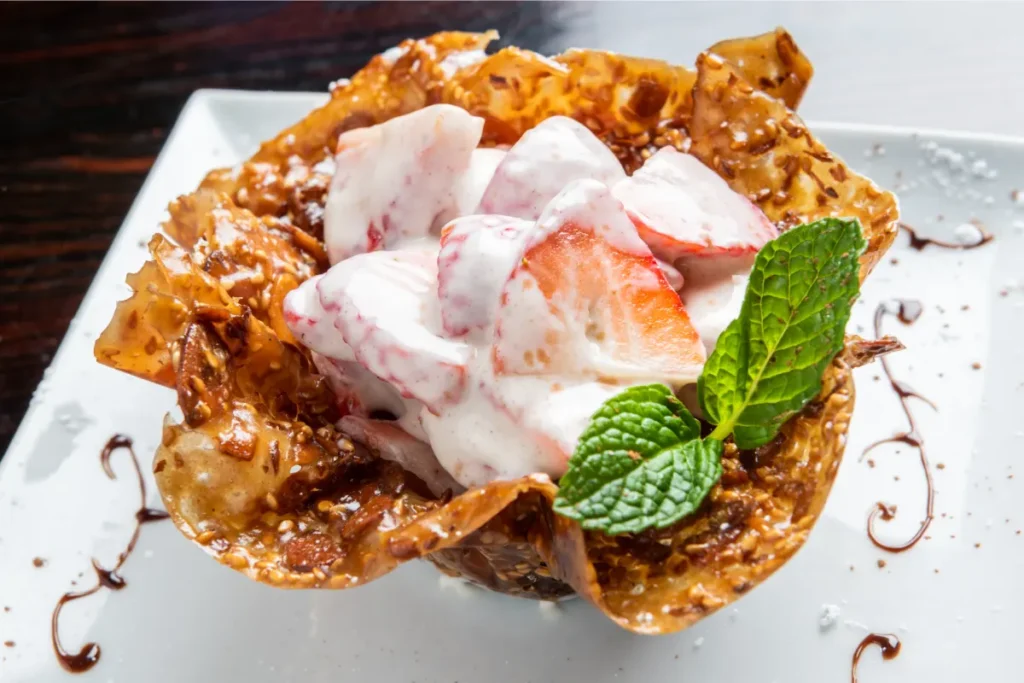 Sliced strawberries and cream on an almond tuile cup garnished with mint, with chocolate drizzle on a white plate.