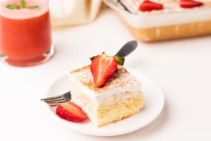 A piece of strawberry tres leches cake garnished with a halved strawberry, dusted with cinnamon, on a white plate with a fork, with more cake and a strawberry smoothie in the background.