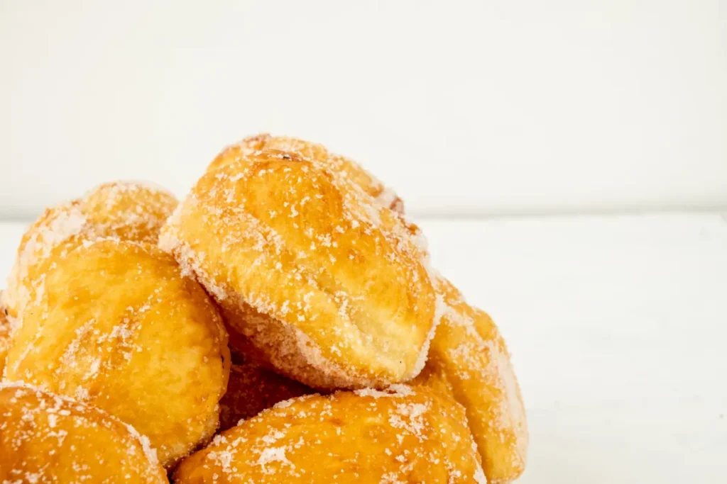 A pile of malasadas heavily dusted with sugar, with a crisp golden-brown exterior, set against a bright white background.