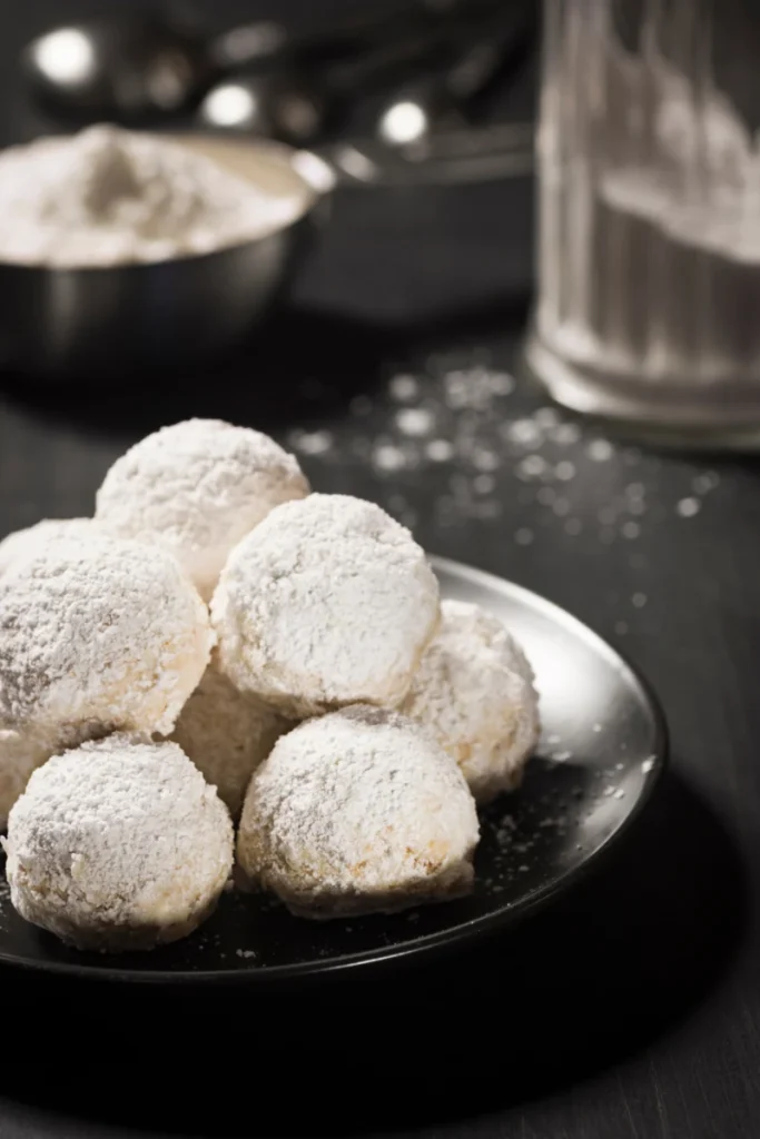 Mexican Wedding Cakes, round cookies heavily dusted with powdered sugar, are centered on a black plate with a blurred kitchen setting in the background, hinting at the baking process.