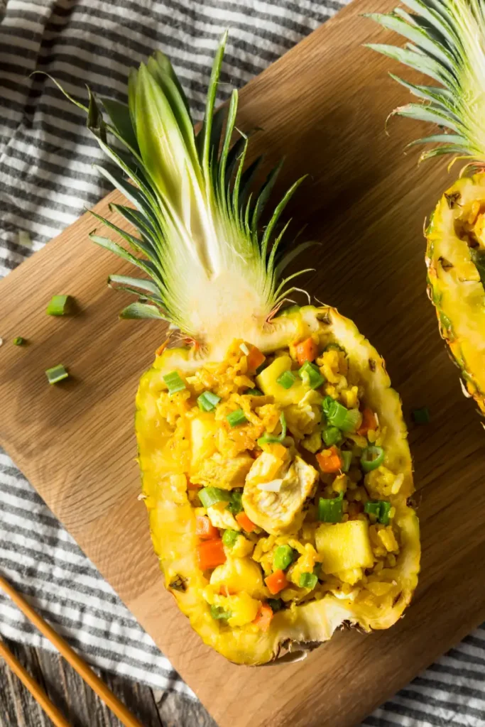 Pineapple fried rice with chunks of pineapple, vegetables, and chicken, served in a pineapple shell on a wooden board, with green onion garnish, over a striped cloth backdrop.