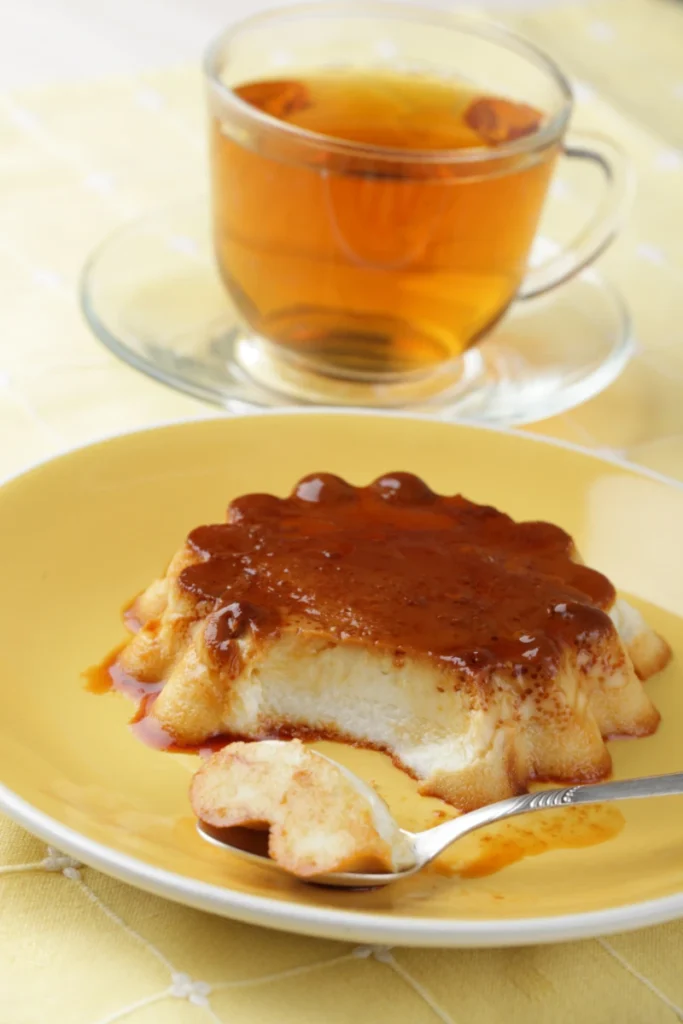 A coconut flan with golden caramel glaze is served on a yellow plate with a fork. In the blurred background, a clear glass cup of tea complements the scene.