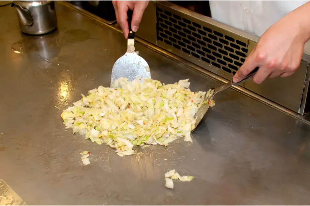 Chef's hands using metal spatulas to chop and mix shredded cabbage on a teppanyaki grill, with a metal teapot in the background.