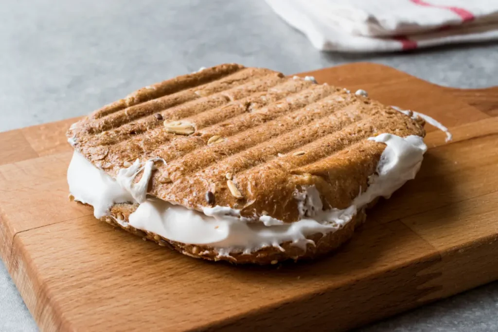 A toasted fluffernutter sandwich with an overflowing marshmallow filling on a wooden board, with a soft-focus background.