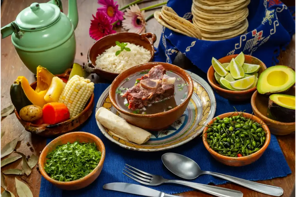 Traditional Caldo de Res served in a clay bowl, surrounded by sides of rice, fresh avocado, lime, a stack of tortillas, and a bowl of chopped greens and peppers, with a green teapot and vibrant flowers in the background.