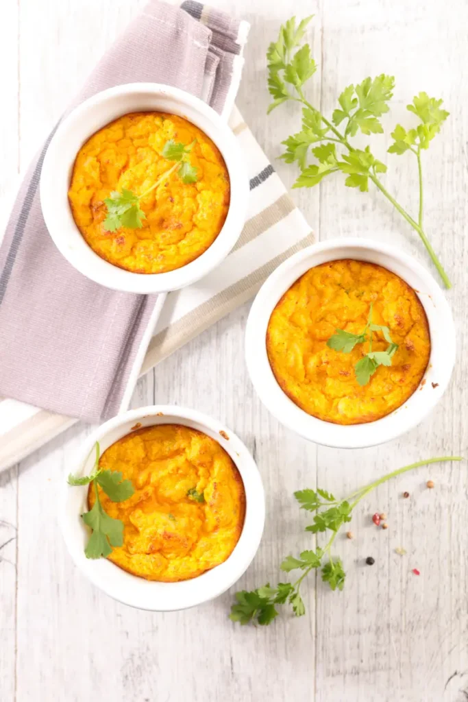 Three individual ramekins of carrot soufflé garnished with fresh parsley on a white wooden table, accompanied by a linen napkin with neutral tones.