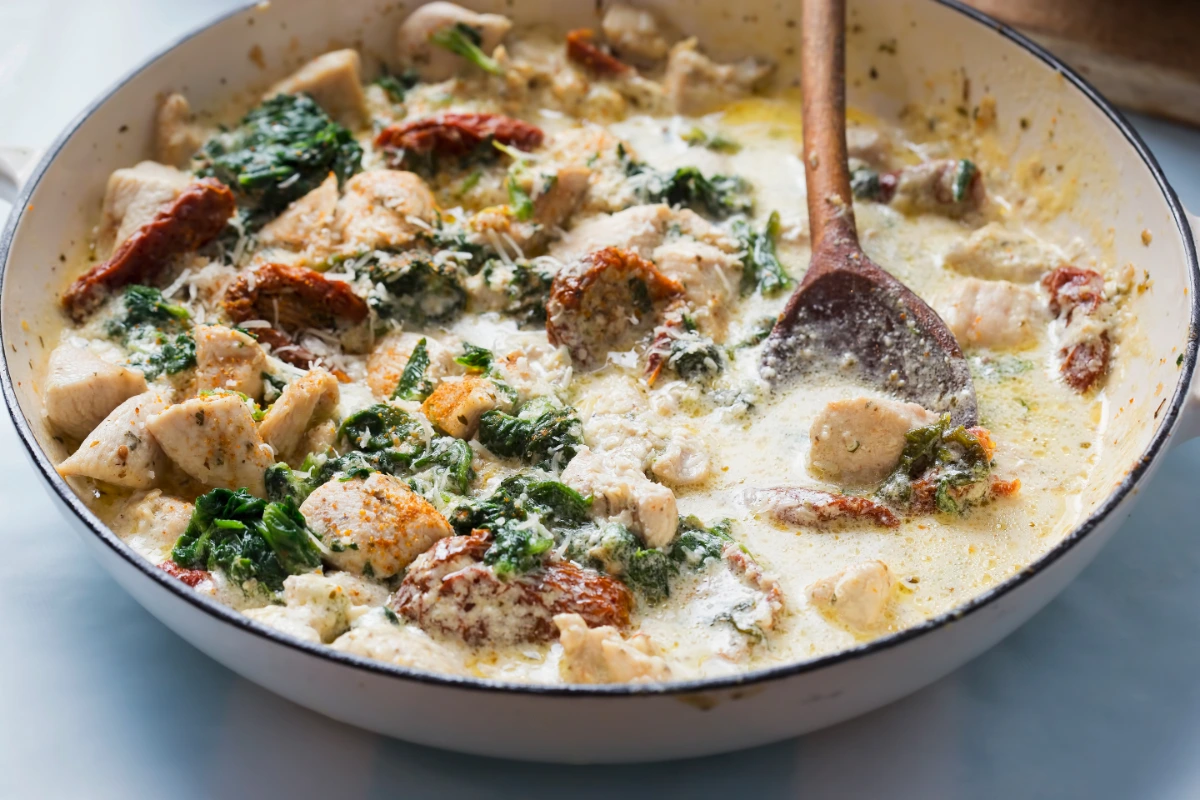 A creamy Tuscan chicken soup with chunks of chicken, sun-dried tomatoes, and spinach, in a white ceramic pot with a wooden spoon.