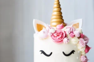 A close-up of a unicorn-themed cake with a golden horn and ears, decorated with pink and cream rosettes, meringues, and a fondant face.