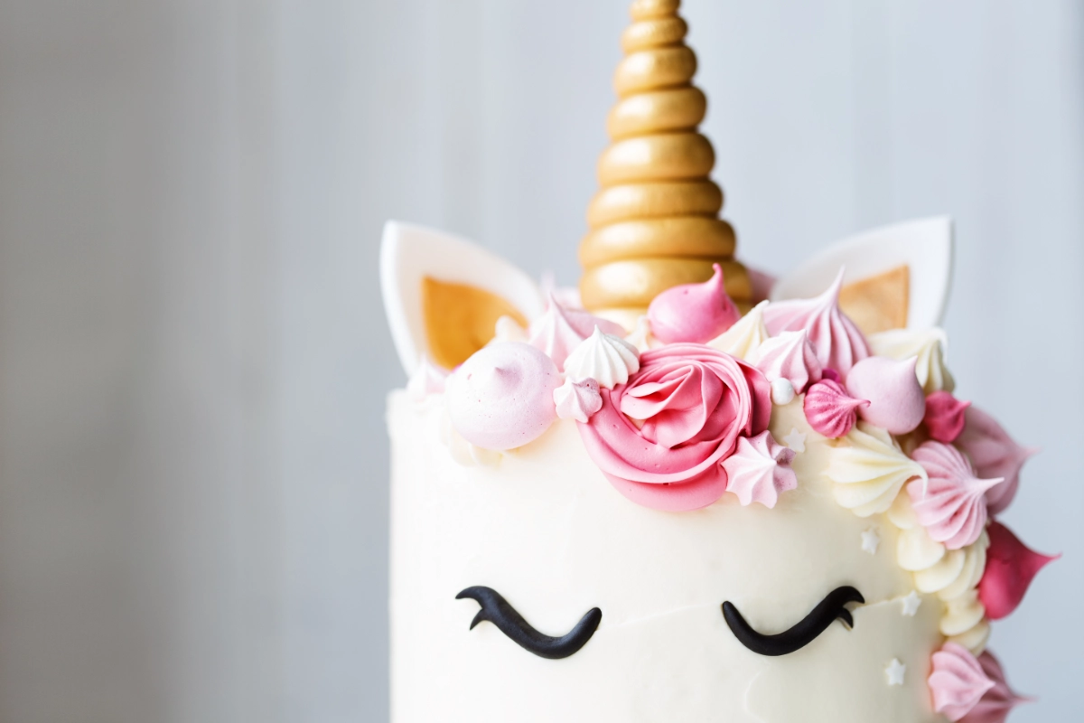 A close-up of a unicorn-themed cake with a golden horn and ears, decorated with pink and cream rosettes, meringues, and a fondant face.