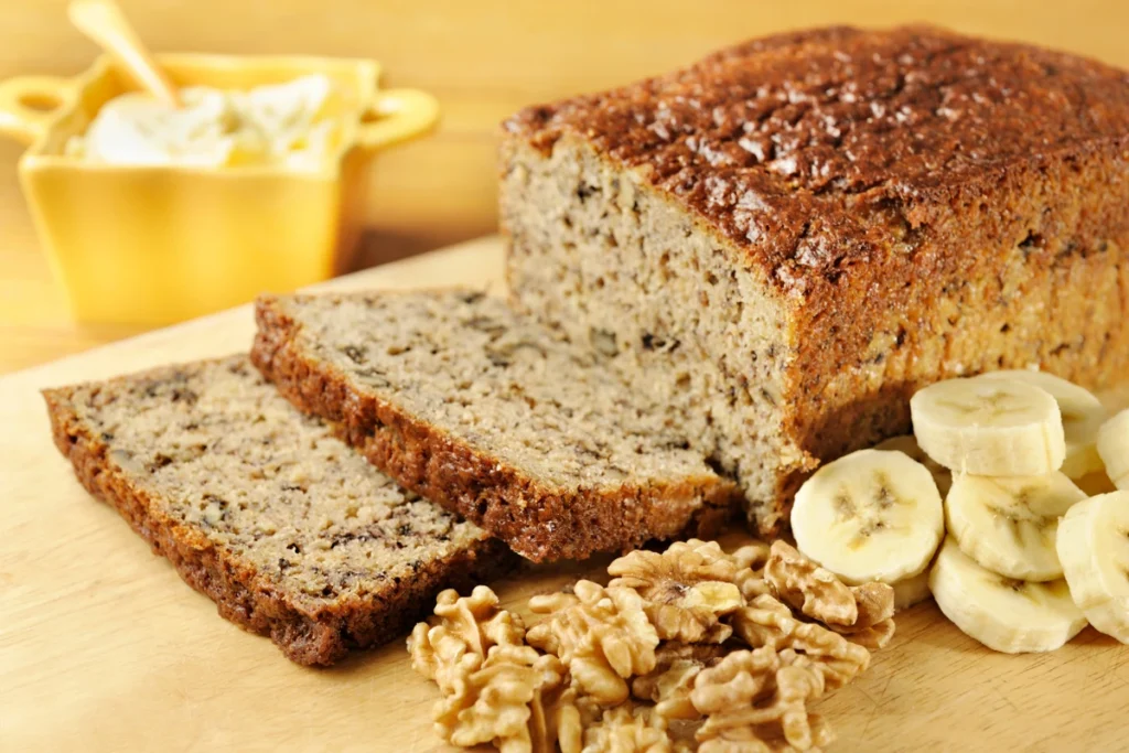 Sliced vegan banana bread with a golden crust, displayed with banana slices and walnuts on a wooden cutting board, and a bowl of butter in the background.