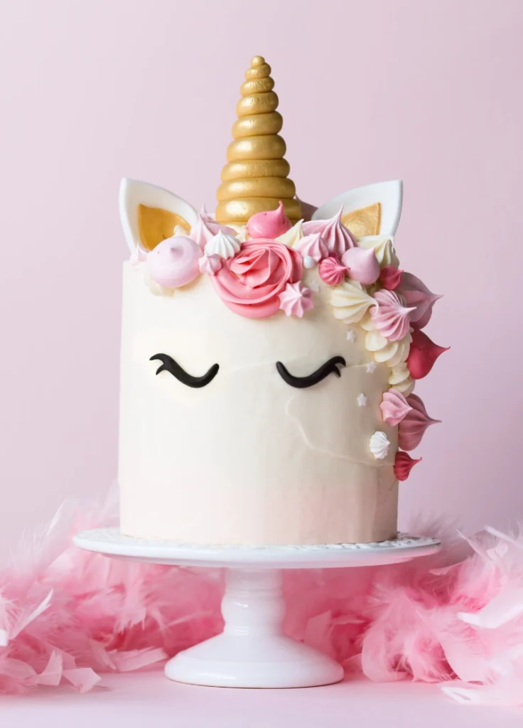 A whimsical unicorn cake with a tall gold horn and fondant ears, adorned with pink flowers and meringues, displayed on a white stand surrounded by soft pink feathers.