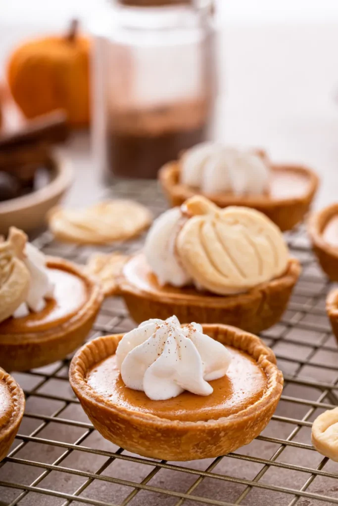 Close-up of mini pumpkin pies topped with whipped cream and a dash of spice, resting on a cooling rack with a blurred background featuring baking ingredients.