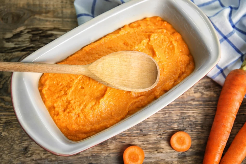 Carrot souffle in a white ceramic dish with a wooden spoon, next to whole carrots and slices on a rustic wooden table, with a blue and white striped cloth partially visible.