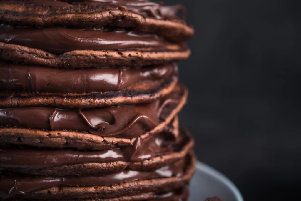 A stack of chocolate pancakes with glossy chocolate spread between the layers, on a dark background.