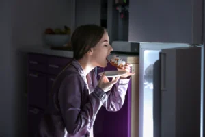 A person in a purple shirt standing by an open refrigerator, biting into a cream-topped pastry on a plate, suggestive of sneaking a late-night snack.