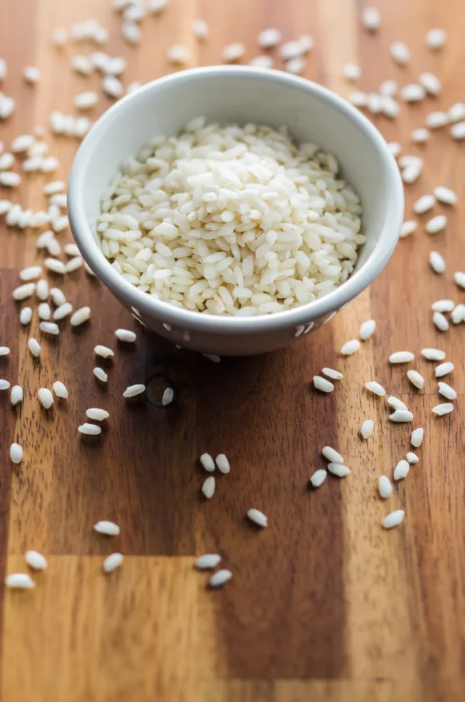 A white bowl filled with uncooked Arborio rice on a wooden surface with scattered grains.