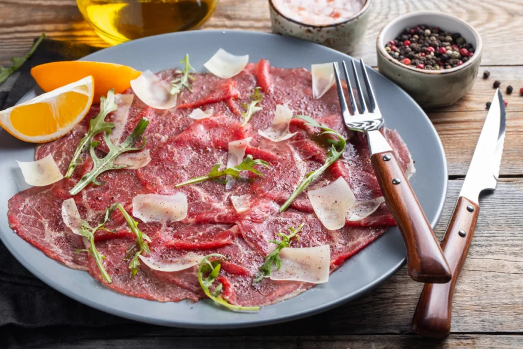 Sliced beef carpaccio garnished with arugula, shaved Parmesan, and orange slices on a gray plate, accompanied by a fork and knife with wooden handles, with olive oil and mixed peppercorns in the background.
