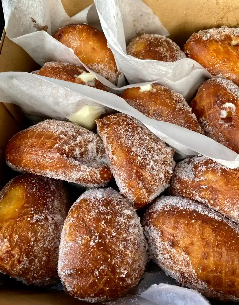 A box of malasadas with golden-brown exteriors, generously coated in granulated sugar, with visible fillings peeking out.