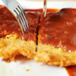 Close-up of a breaded cutlet covered in katsu sauce, with a fork cutting into it on a white plate.