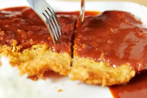 Close-up of a breaded cutlet covered in katsu sauce, with a fork cutting into it on a white plate.