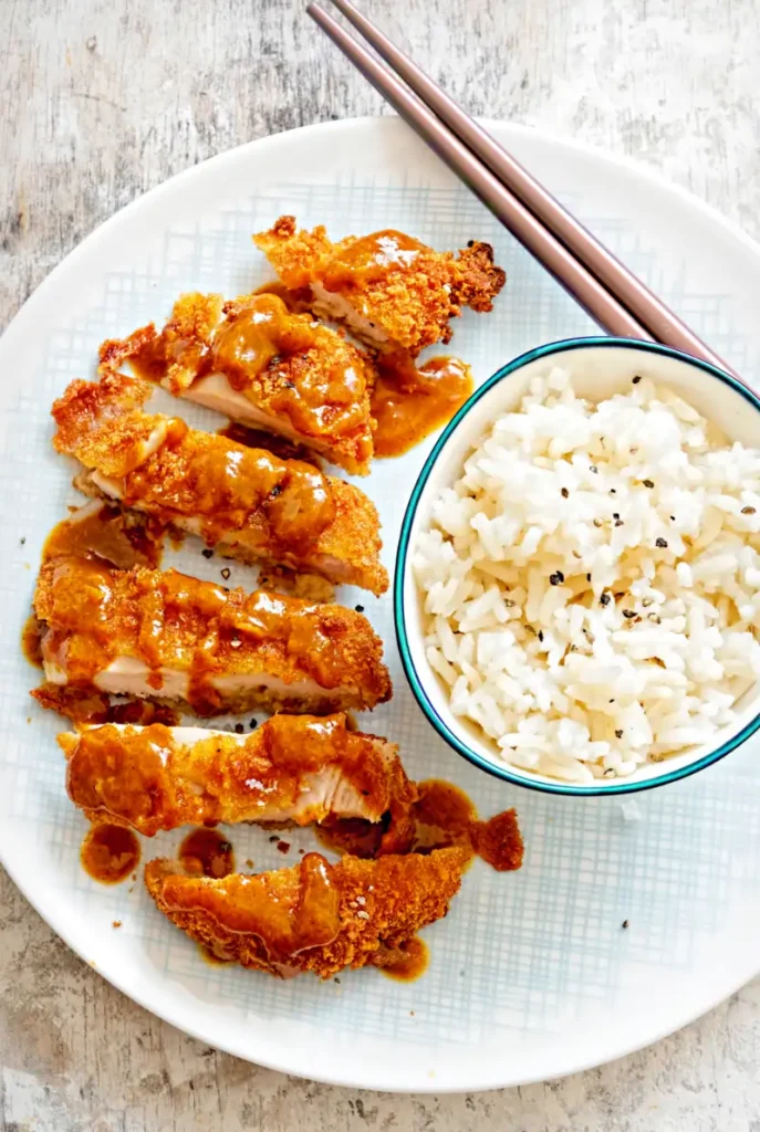 Sliced breaded cutlet topped with katsu sauce served next to a bowl of white rice on a round plate with chopsticks.