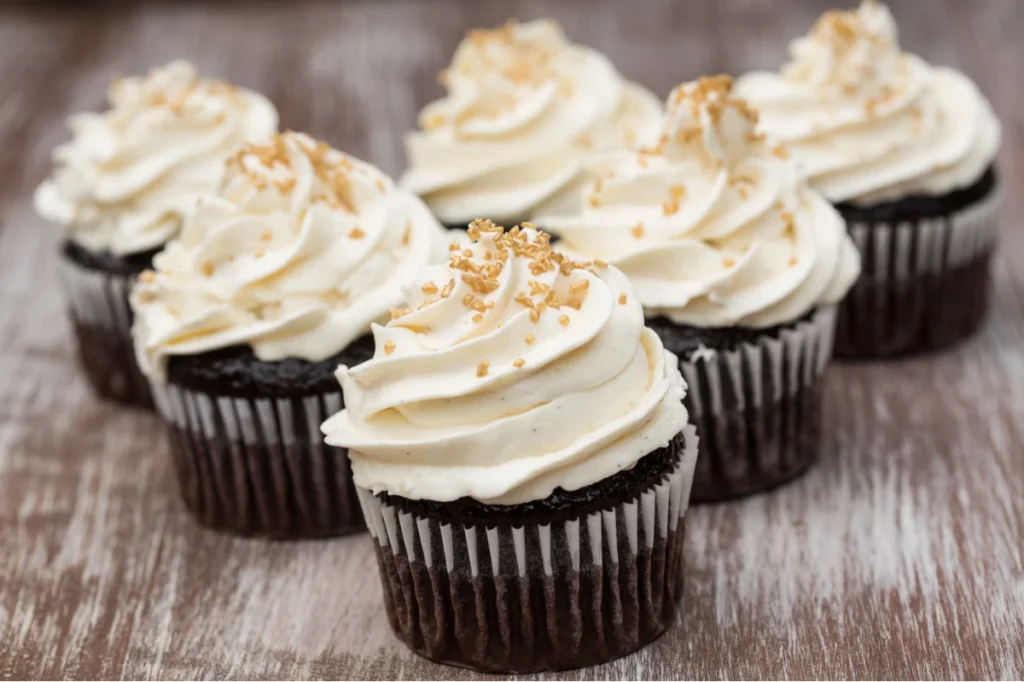 Close-up of five chocolate cupcakes topped with swirls of Russian buttercream and sprinkled with golden sugar crystals, on a wooden surface.