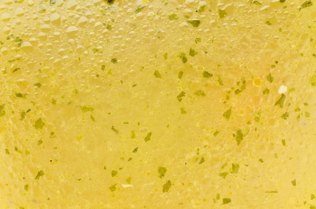 Close-up view of chicken bouillon showing a yellow, translucent liquid speckled with green herbs and tiny bubbles.