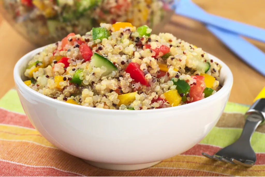 Close-up view of a zesty quinoa salad in a white ceramic bowl. The salad features a colorful mix of red and yellow bell peppers, black quinoa grains, and cucumber pieces. The bowl is placed on a multi-colored striped napkin, with a serving spoon nearby, set against a wooden table background.