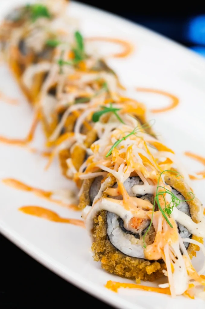 Close-up of crunchy roll sushi topped with spicy mayo and shredded cheese, garnished with delicate green herbs, on a long white plate with orange sauce drizzle.