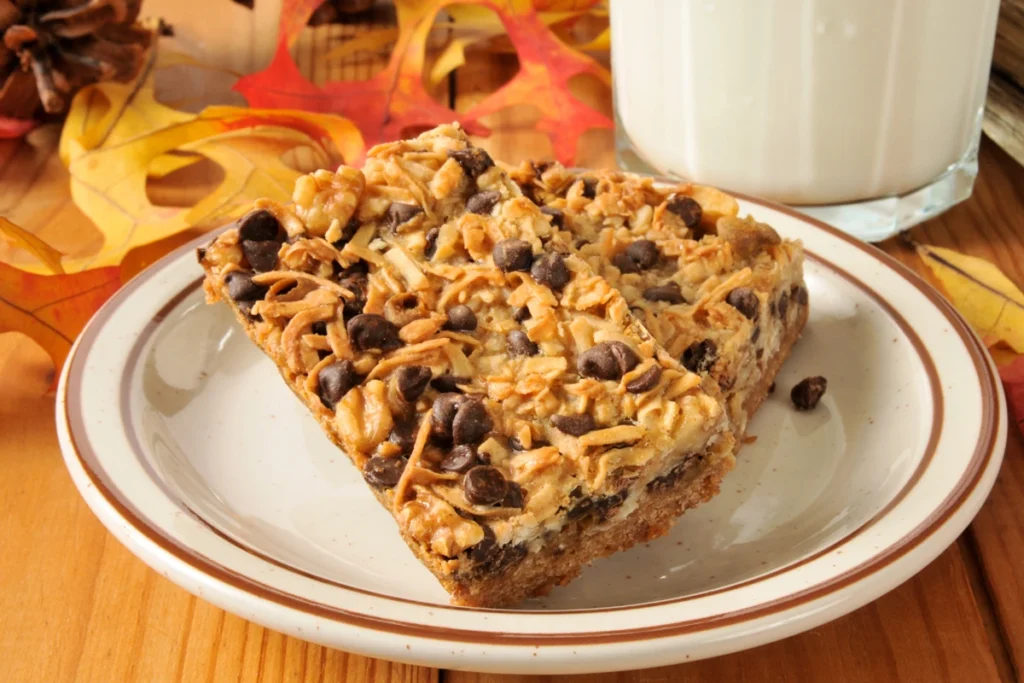 A close-up view of a 7 layer bar on a ceramic plate, garnished with chocolate chips and coconut shavings. The bar is set against a backdrop of autumn leaves and a glass of milk.