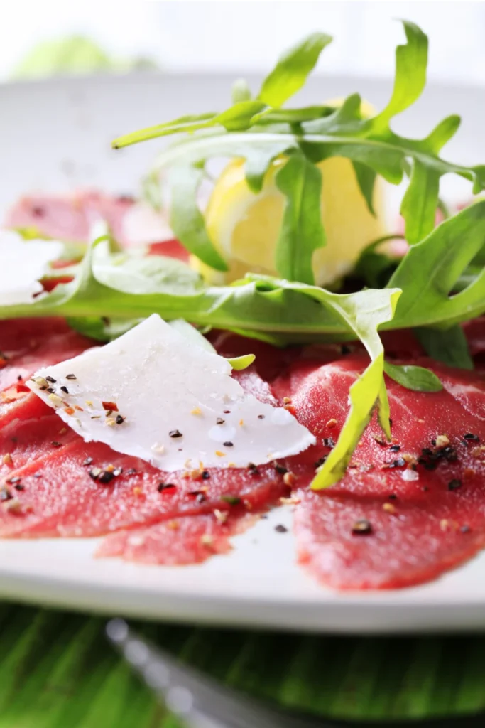 Close-up view of beef carpaccio topped with arugula, a lemon wedge, and Parmesan shavings on a white plate, with a focus on the fresh ingredients and seasoning.